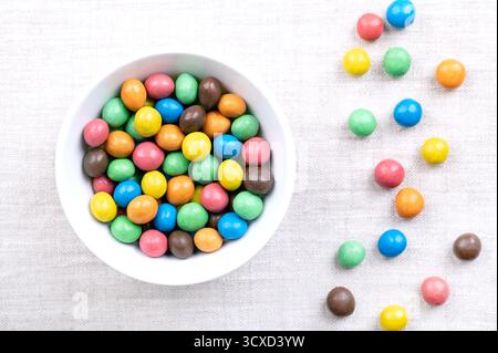 Colorful sugar-coated chocolate dragees, in a white bowl, on linen. Dragee chocolate confectionery, consisting of a multi colored candy shell. Stock Photo