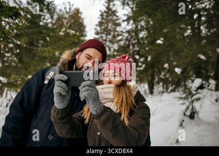 Young woman and man in outwear hiking together in winter time and taking selfie in evergreen forest Stock Photo