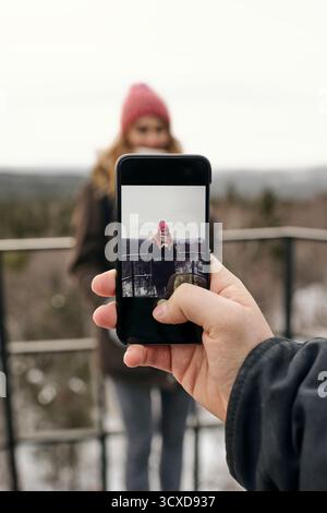 Crop male hand using smartphone and photographing smiling girlfriend on terrace against highlands Stock Photo
