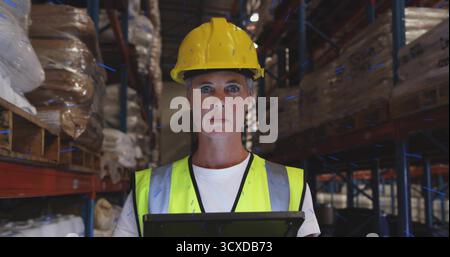 Inspecting inventory, supervisor wearing hard hat and vest holding tablet in aisle among pallets Stock Photo