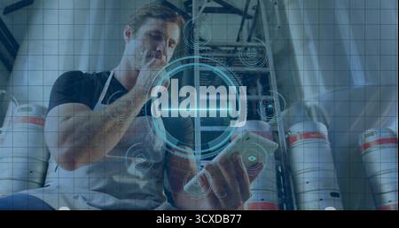 Holding smartphone, touching chin, worker in grey apron in brewery storeroom with kegs and tanks Stock Photo