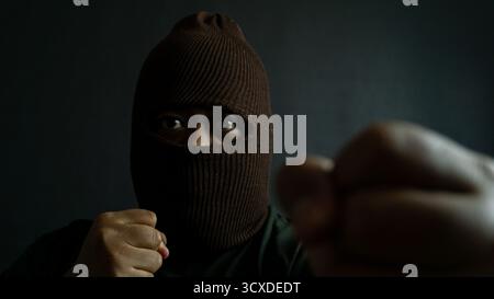 Masked man in brown cloth mask striking a fighting pose, clenched fist raised with eyes visible, intense action mood on dark background Stock Photo