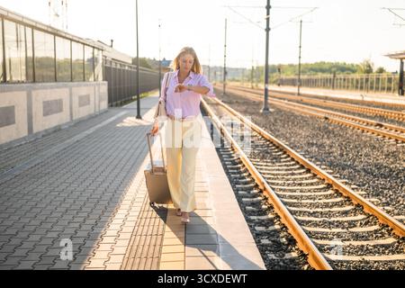 Confident and elegant businesswoman with suitcase looking at her wristwatch while walking at the railway station. Stock Photo