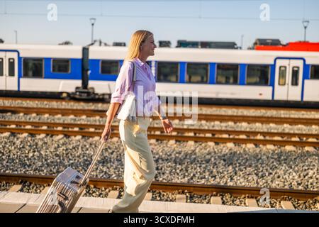 Elegant businesswoman walking confidently with suitcase at the sunny railway station. Stock Photo
