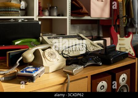 Vintage electronics including rotary phone, typewriter, retro video game console, and audio equipment displayed on wooden counter in thrift shop setting with shelves in background Stock Photo