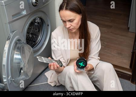 Woman sits near a washing machine, holding a limescale-covered heating element and a pump motor, looking puzzled while diagnosing or repairing the hou Stock Photo