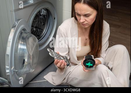 Woman sits near a washing machine, holding a limescale-covered heating element and a pump motor, looking puzzled while diagnosing or repairing the hou Stock Photo