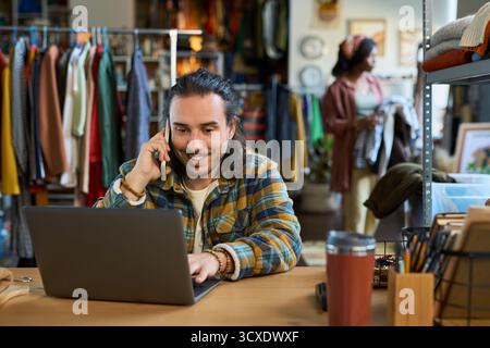 Caucasian young adult man sitting at desk smiling while talking on smartphone and using laptop in thrift shop with racks of clothing in background, African American young adult woman browsing clothes Stock Photo