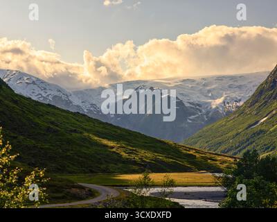 The dramatic, snow-covered glacier and peaks of a vast valley are illuminated by warm, late-afternoon sun cutting through clouds, overlooking a lush g Stock Photo