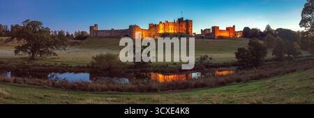 A view at dusk in early Autumn of floodlit Alnwick Castle in Alnwick, Northumberland viewed from the Pastures looking over the River Aln Stock Photo