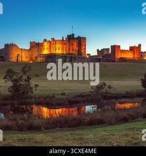 A view at dusk in early Autumn of floodlit Alnwick Castle in Alnwick, Northumberland viewed from the Pastures looking over the River Aln Stock Photo