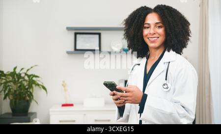 Confident Female Doctor With Stethoscope Using Smartphone In Modern Clinic Setting For Telemedicine And Patient Care Stock Photo