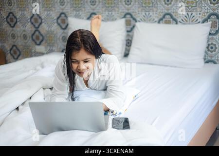 Asian woman using laptop on bed at home, relaxing and working remotely, modern lifestyle and technology concept, comfortable workspace with casual moo Stock Photo