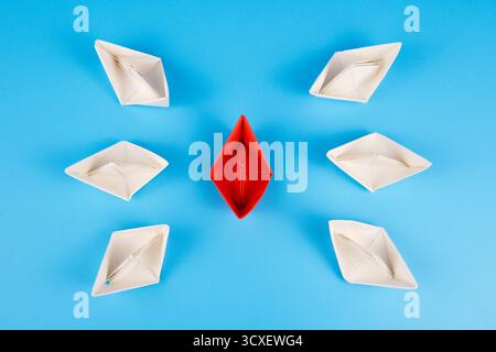 Symbolic photo showing a red paper boat surrounded by white boats on blue background, representing humanitarian aid ship surrounded. Stock Photo