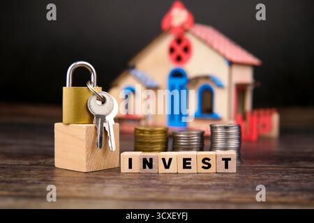 Padlock and keys placed on wooden block beside stacked coins and wooden cubes spelling “INVEST,” with a miniature house in the background. Stock Photo