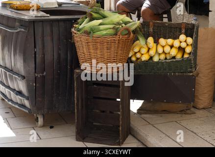 A rustic street vendor display features baskets piled high with fresh corn on the cob, both husked and unhusked, set atop a dark wooden crate. Stock Photo