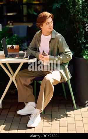A young man sits at a cafe table, focused on his laptop while taking in the lively setting. Stock Photo