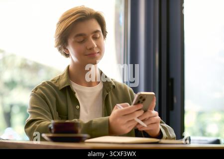 A young man sits at a cafe, focused on his smartphone while enjoying a warm beverage. Stock Photo