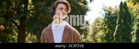 A young man strolls through a vibrant autumn park, dressed in a stylish coat and headphones, banner Stock Photo