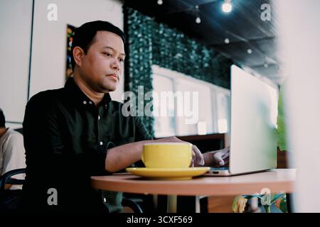 Young Asian Man Working with Laptop in Coffee Shop While Drinking Coffee Stock Photo