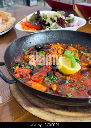 Tasty and filling seafood stew made with shrimp, mussels, clams and squid in a rich tomato broth. The dish with herbs and lemon in a cast iron skillet Stock Photo