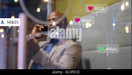 Talking businessman in suit holding smartphone in lounge, with pendant lights and social icons Stock Photo