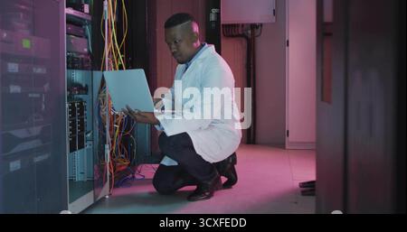 Kneeling IT technician wearing lab coat typing on laptop in server room, with Ethernet cables Stock Photo