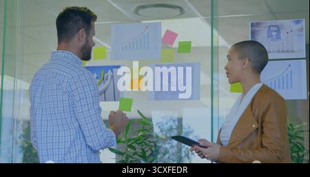 Office professionals analyzing charts on glass partition in office, with sticky notes and tablet Stock Photo