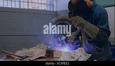 Welding technician directing torch at car body in workshop, wearing helmet, with sparks, copy space Stock Photo