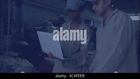 Collaborating two technicians in coveralls reviewing inventory on laptop in warehouse, with pallets Stock Photo
