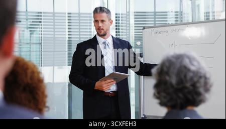 Presenting suited businessman holding tablet, gesturing to whiteboard in meeting room, with charts Stock Photo