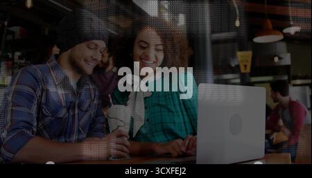 Smiling pair wearing beanie and blouse viewing silver laptop at cafe bar, with cream-topped shake Stock Photo
