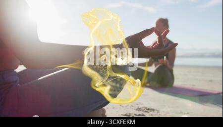 Holding meditation mudra, woman in leggings and top sitting cross-legged on beach, with yoga mats Stock Photo