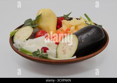 Fresh vegetables including eggplant, potato, onion, carrot, tomato, and herbs arranged in a clay plate on white background. Concept of healthy food, c Stock Photo