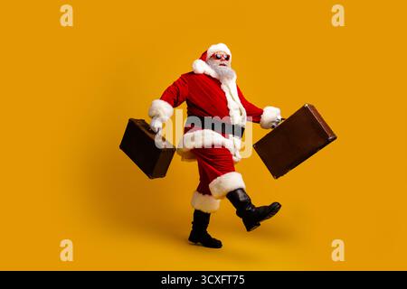 Santa Claus with suitcases striding confidently against a yellow background conveying holiday spirit and travel fun Stock Photo