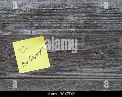 Yellow sticky note with handwritten "Be Happy!!" and smiley on rustic wood background Stock Photo