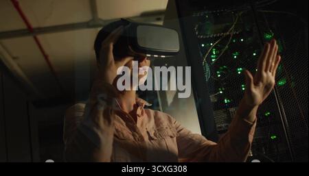 Interacting IT tech wearing shirt with VR headset in data center corridor, with blinking rack LEDs Stock Photo