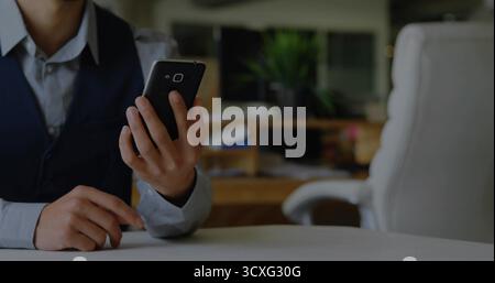 Holding smartphone, man in dress shirt and vest tapping screen at round white table in workspace Stock Photo