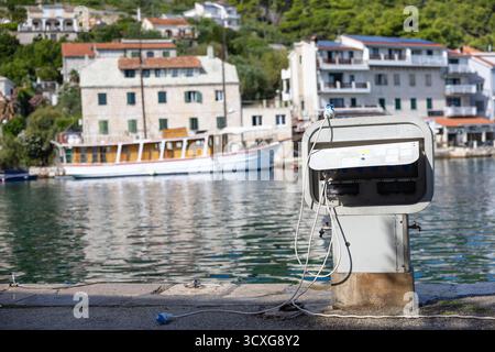 A seaside harbor scene shows a weathered charging station on a concrete pier, calm water, white boats, and colorful townhouses set against lush green Stock Photo