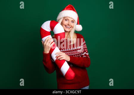 Cheerful woman wearing Santa hat holding candy cane on green background enjoying festive Christmas holiday vibes Stock Photo