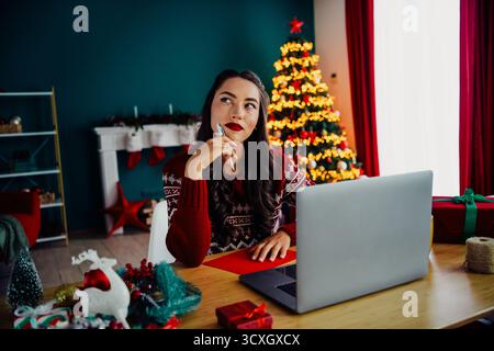 Festive woman in cozy sweater working on laptop amidst Christmas decorations and tree in a beautifully lit room Stock Photo