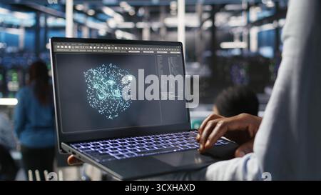 Close up of data center worker using laptop to interpret AI generated data analytics graphs. Server room technician using artificial intelligence on notebook displaying system metrics, camera A Stock Photo