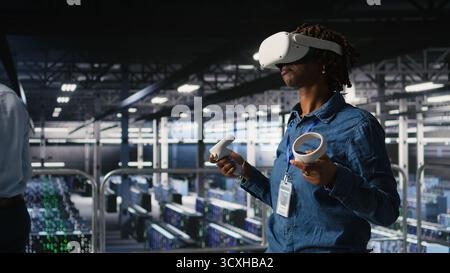 Data center programmer using virtual reality technology to simulate artificial intelligence neural networks. Woman in server farm using VR headset equipment to oversee AI systems, Stock Photo