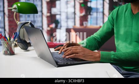 Freelancer creating articles content for his online blog at home desk, writing and working on storytelling with his laptop. Black guy composes social media posts, multitasking. Camera B. Stock Photo
