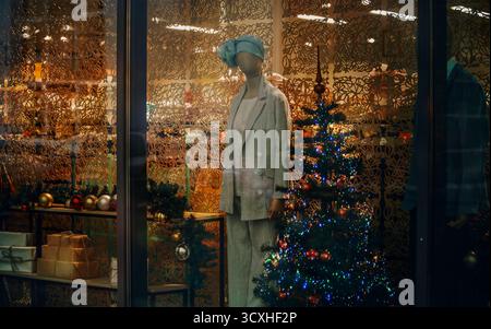 Festive shop window with a mannequin in a stylish suit, decorated with a Christmas tree, ornaments, and gift boxes in warm cozy lighting. Stock Photo