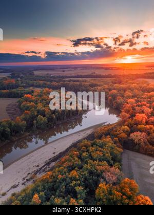 A warm October sunset behind a bend in the river. Red, orange, and yellow trees adorn the river banks. There are clouds on the horizon and faint rays. Stock Photo