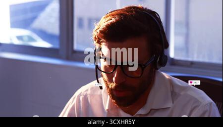 Bearded man wearing headset and eyeglasses reading customer message at office desk near window Stock Photo