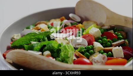 White ceramic bowl resting on light tabletop, with salad, tomatoes, croutons, wooden tongs Stock Photo