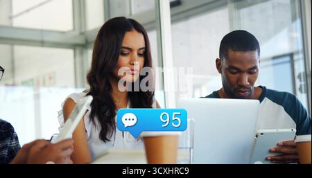 Collaborating pair in casual attire focusing on devices at office table, with notification bubble Stock Photo