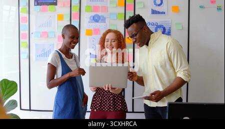Collaborating three colleagues discussing data on laptop and tablet in meeting space with notes Stock Photo